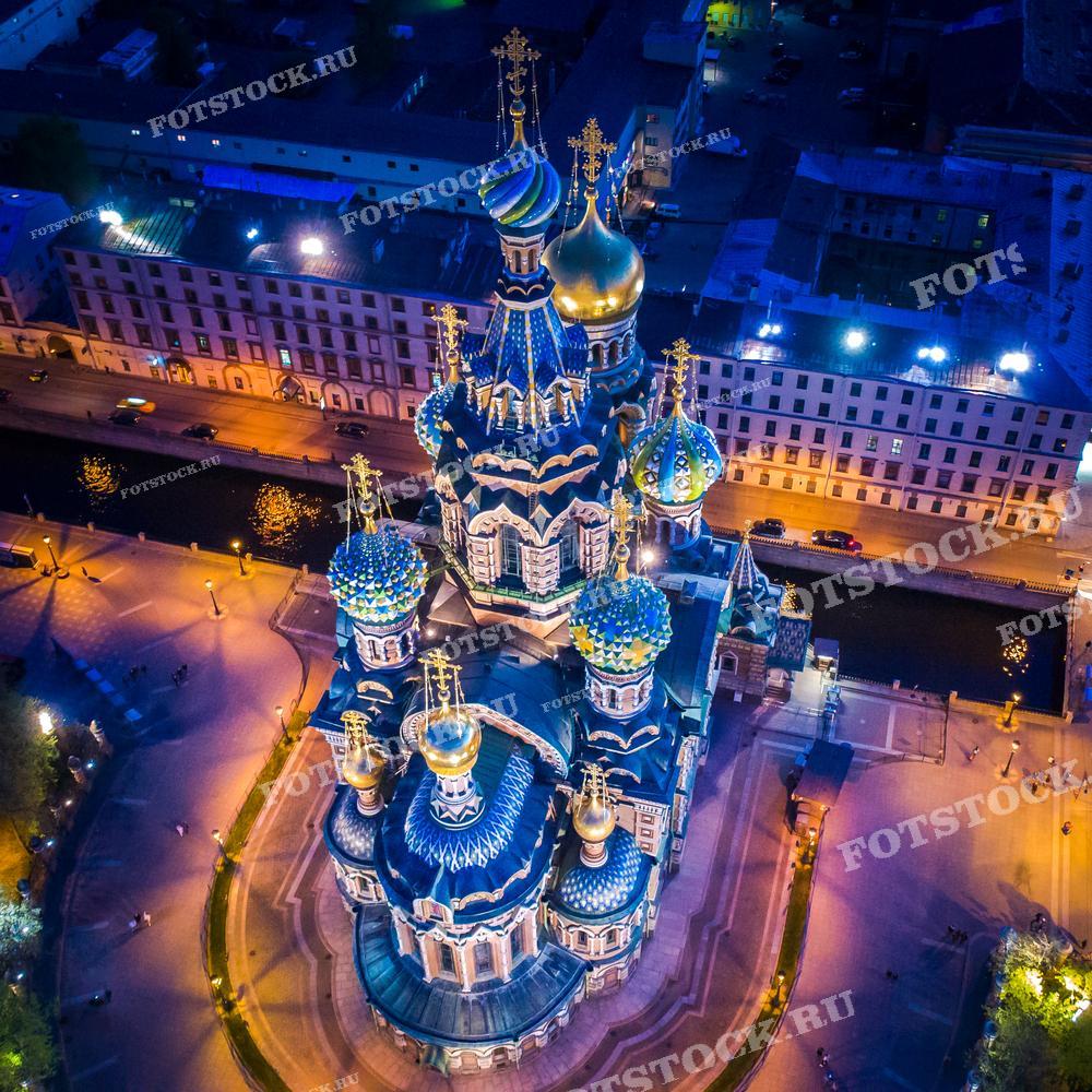 На фотографии изображён Храм Спаса-на-Крови в Санкт-Петербурге, снятый с высоты в вечернее время. Храм освещен огнями, что подчеркивает его красочные купола и детали архитектуры. Здание окружено городскими улицами и каналами, создавая атмосферу исторического центра города. Это изображение может быть использовано в туристических путеводителях, рекламных материалах, посвящённых Санкт-Петербургу, а также в образовательных и культурных публикациях.